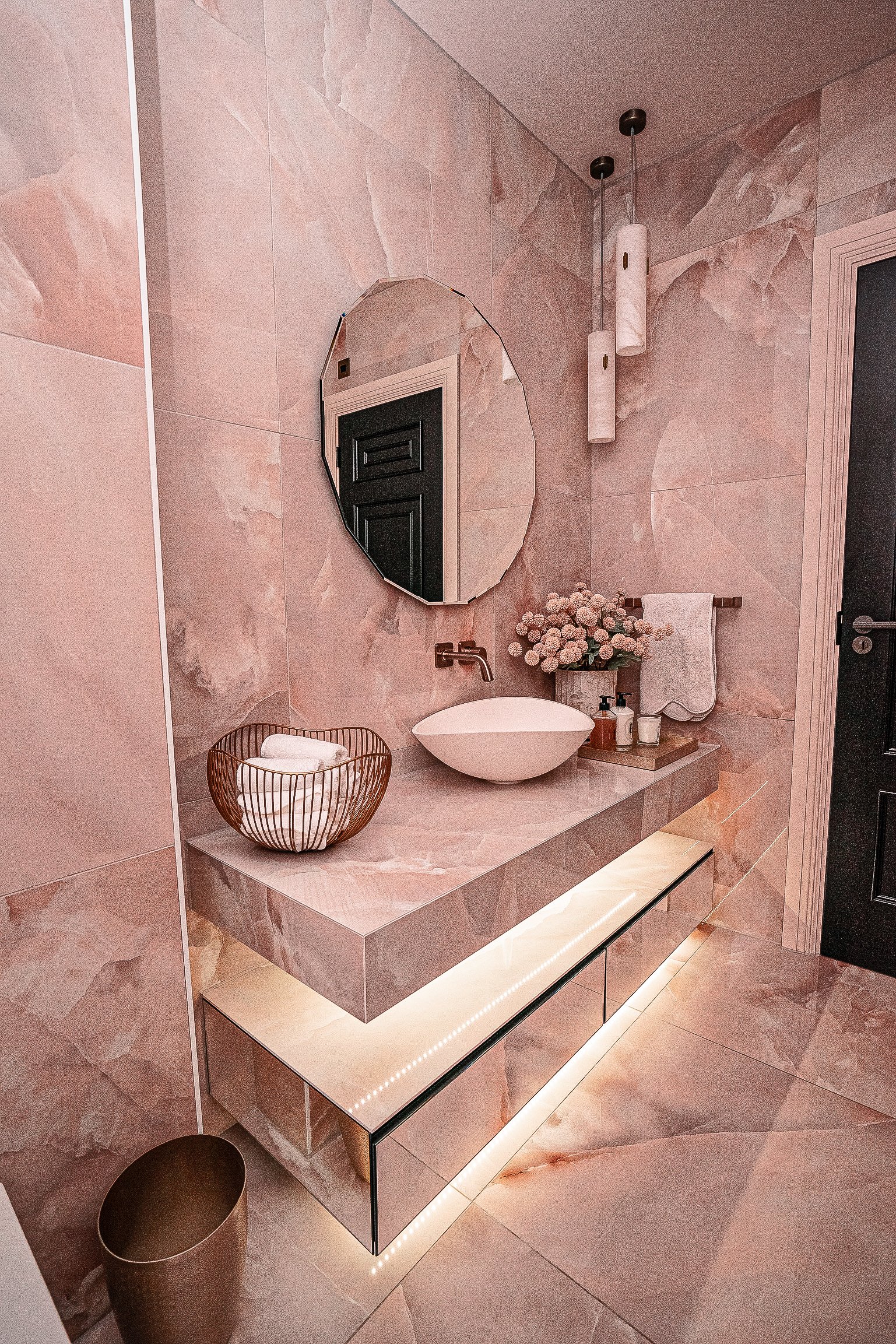 Modern bathroom with marble tiles, round mirror, round vessel sink, wire basket with towels, decorative flowers, pendant lights, and LED-lit vanity.