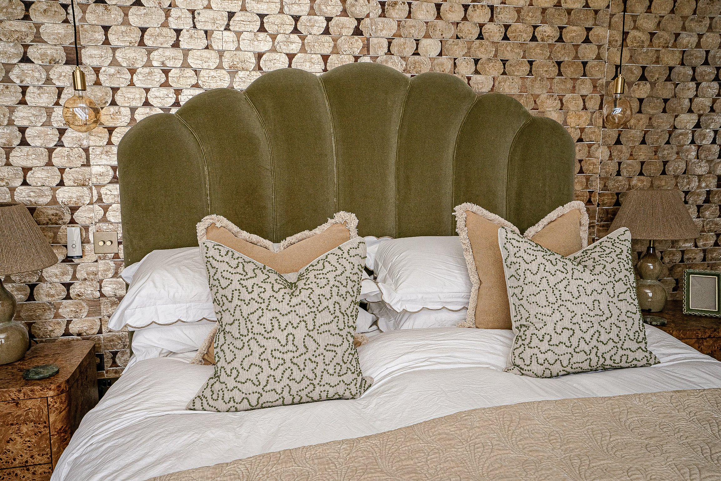 Bedroom with green velvet headboard, patterned pillows, beige quilt, wooden bedside tables with lamps, and a textured wall background.