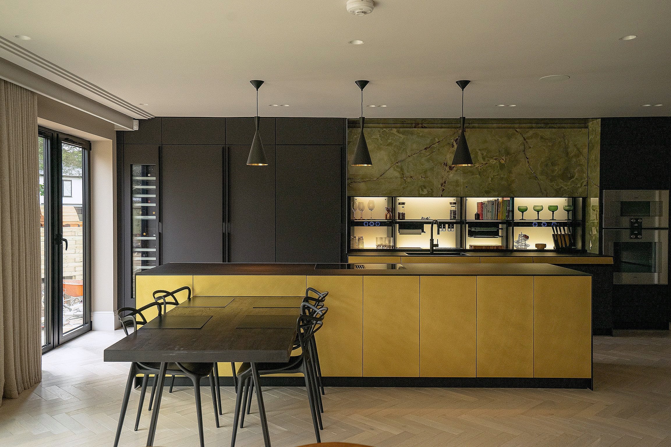 Modern kitchen interior with large island, black cabinets, and dining table with chairs, featuring pendant lighting and glass doors.