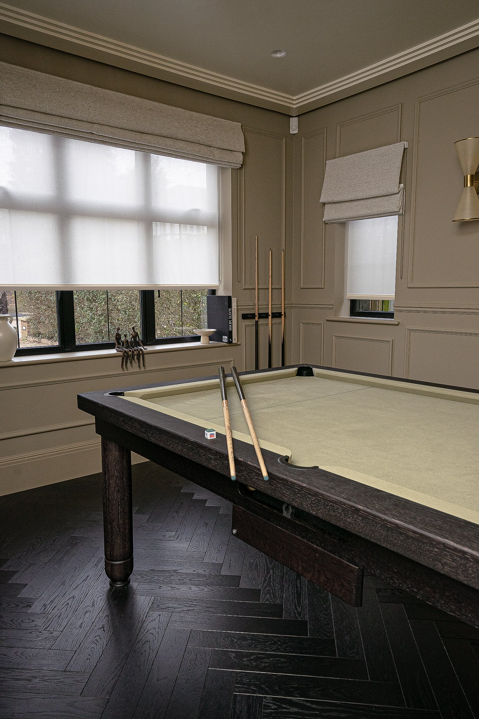Elegant billiards room with a pool table, two pool cues on the table, three cues in a stand, a window with white blinds, and stylish decor.