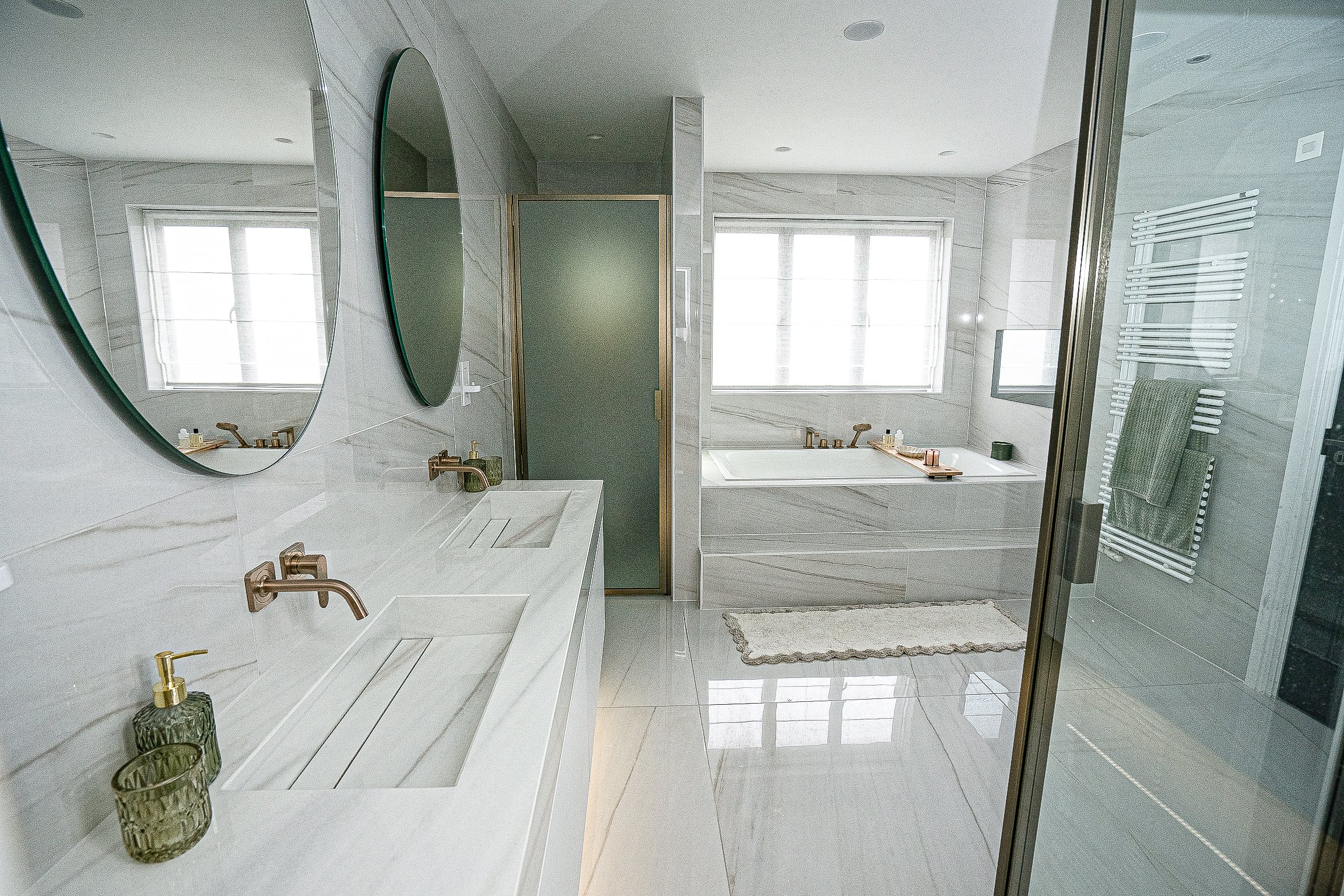 Modern bathroom with dual sinks, circular mirrors, large bathtub, and glass-enclosed shower; light marble surfaces and gold fixtures.