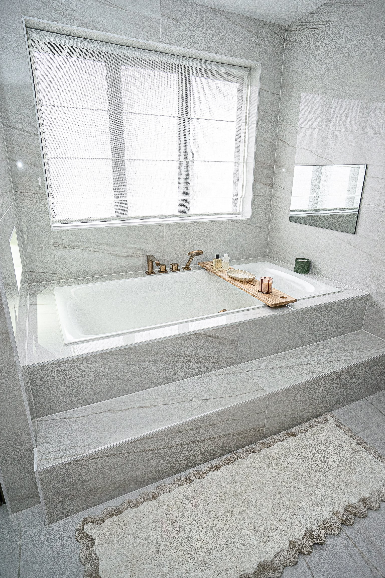 Modern bathroom with white tub, marble tiles, and a wooden bath tray holding toiletries and a candle, near a window and a bath mat.