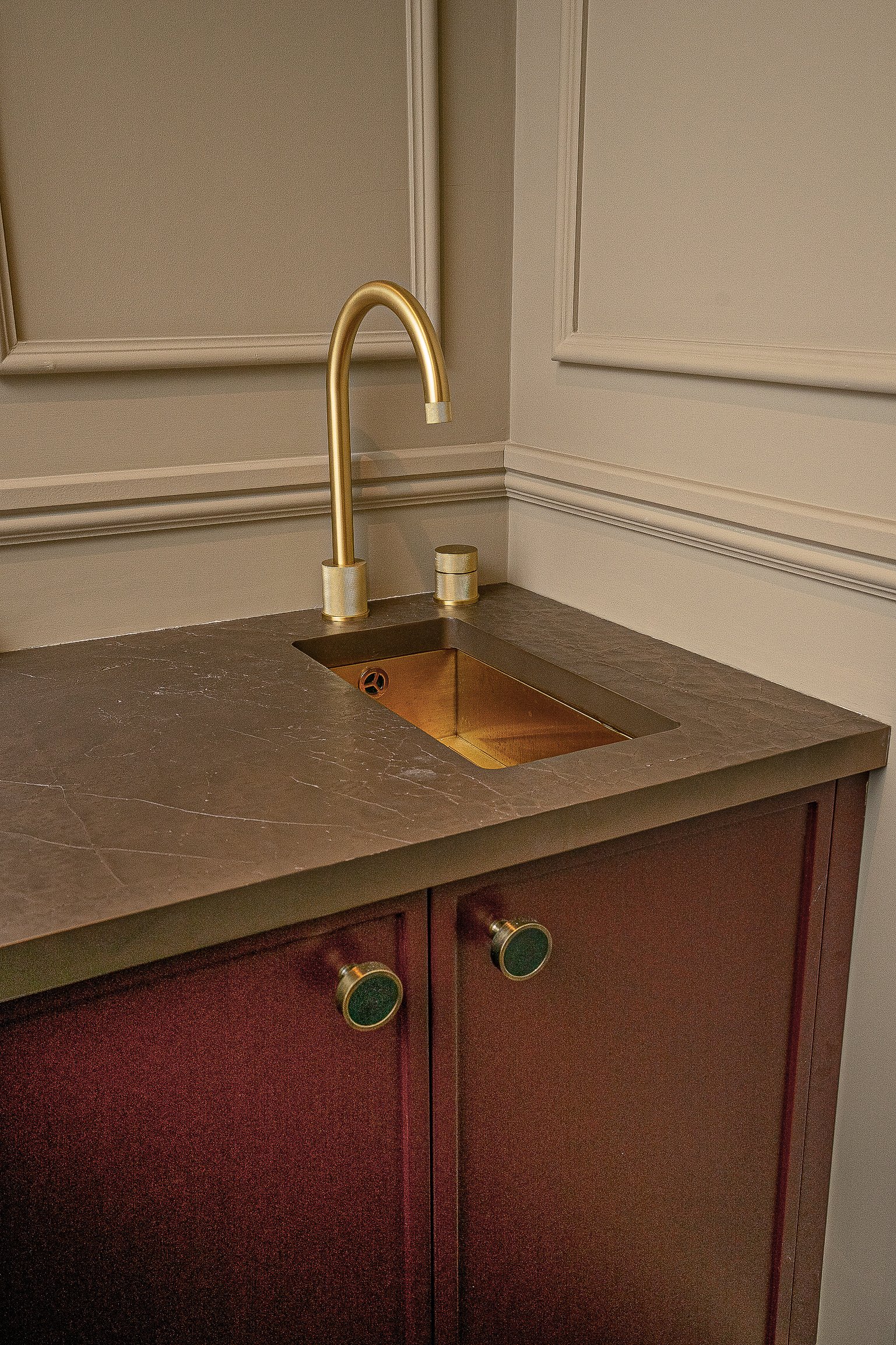 Elegant bathroom sink with a gold faucet, gold basin, and brown countertop, set against a beige paneled wall, above maroon cabinets with decorative handles.