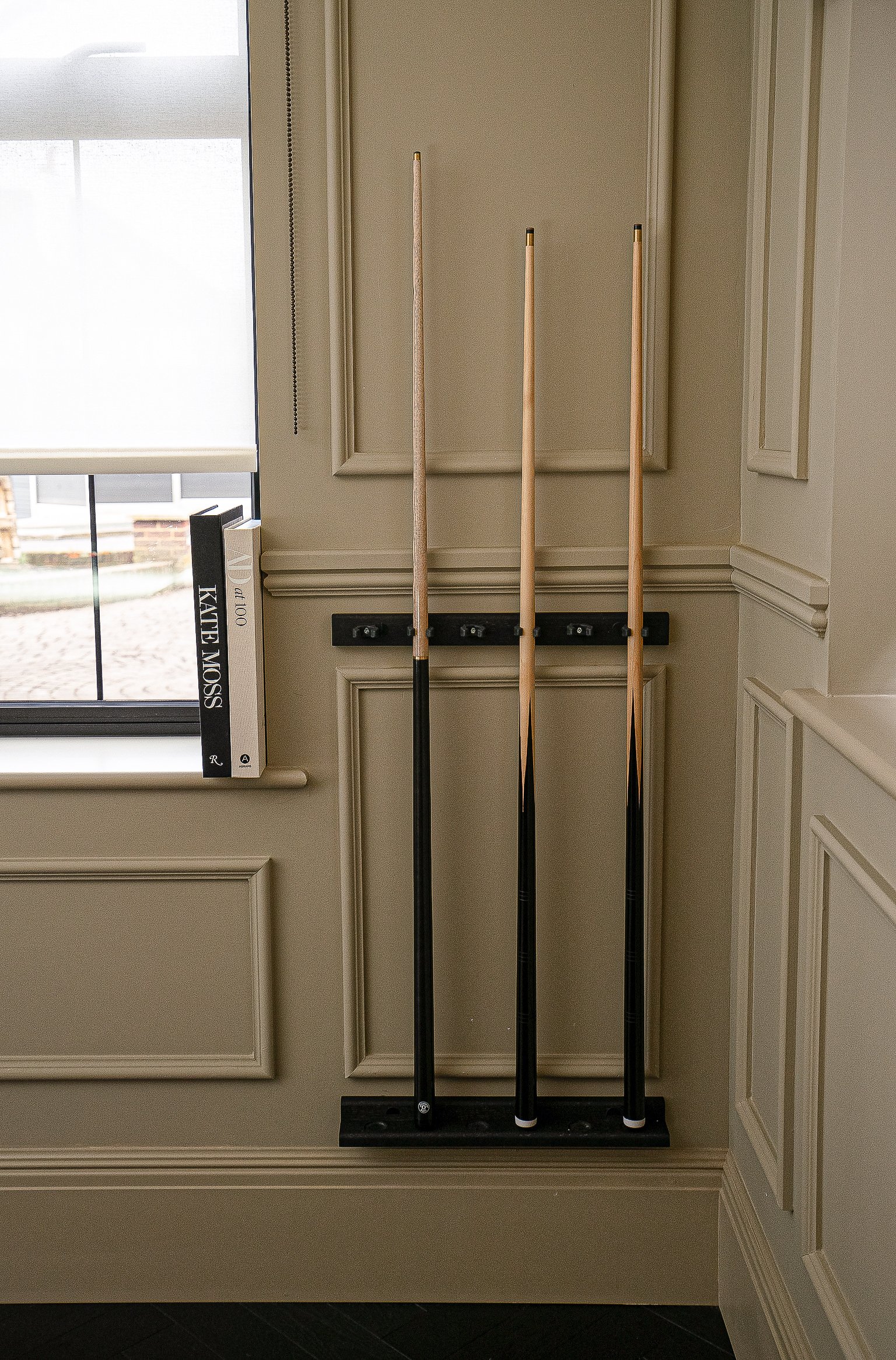 Three pool cues in a wall-mounted rack next to a window with a book titled 'Kate Moss' standing on the windowsill.