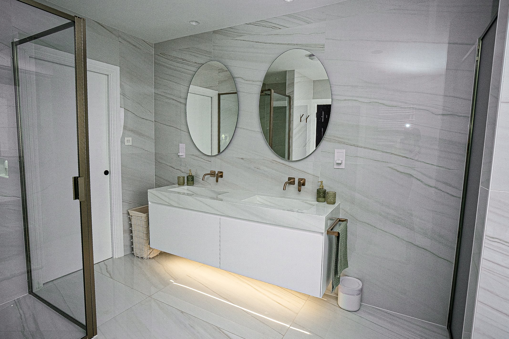 Modern bathroom with dual floating sinks, oval mirrors, and marble tiles. Features glass shower door and wall-mounted faucets.
