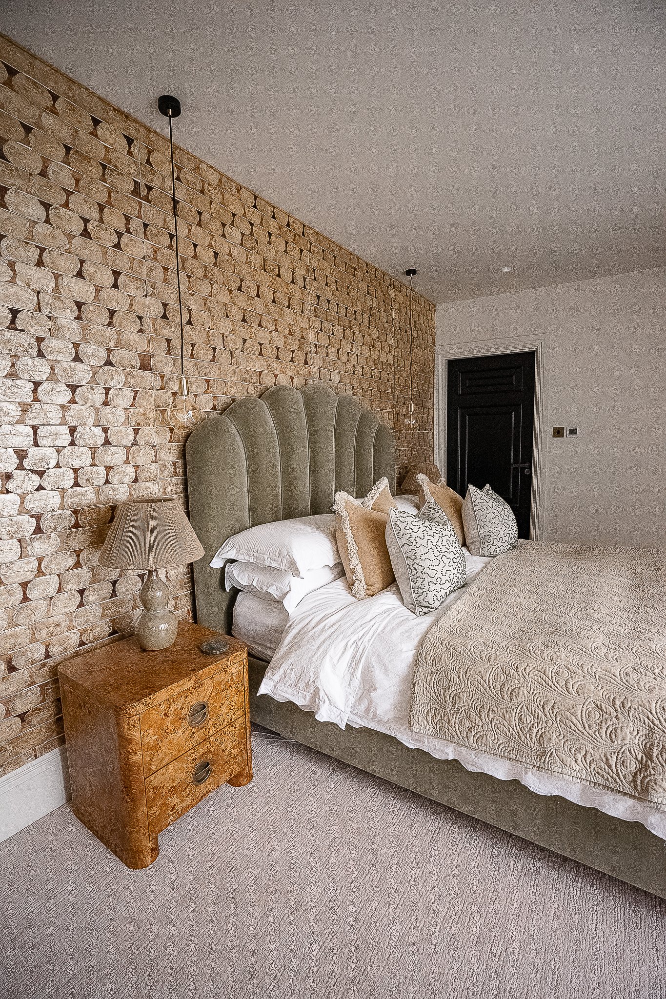 Modern bedroom with textured wall, green velvet headboard, beige bedding, and decorative pillows. Wooden nightstand with a lamp beside the bed.