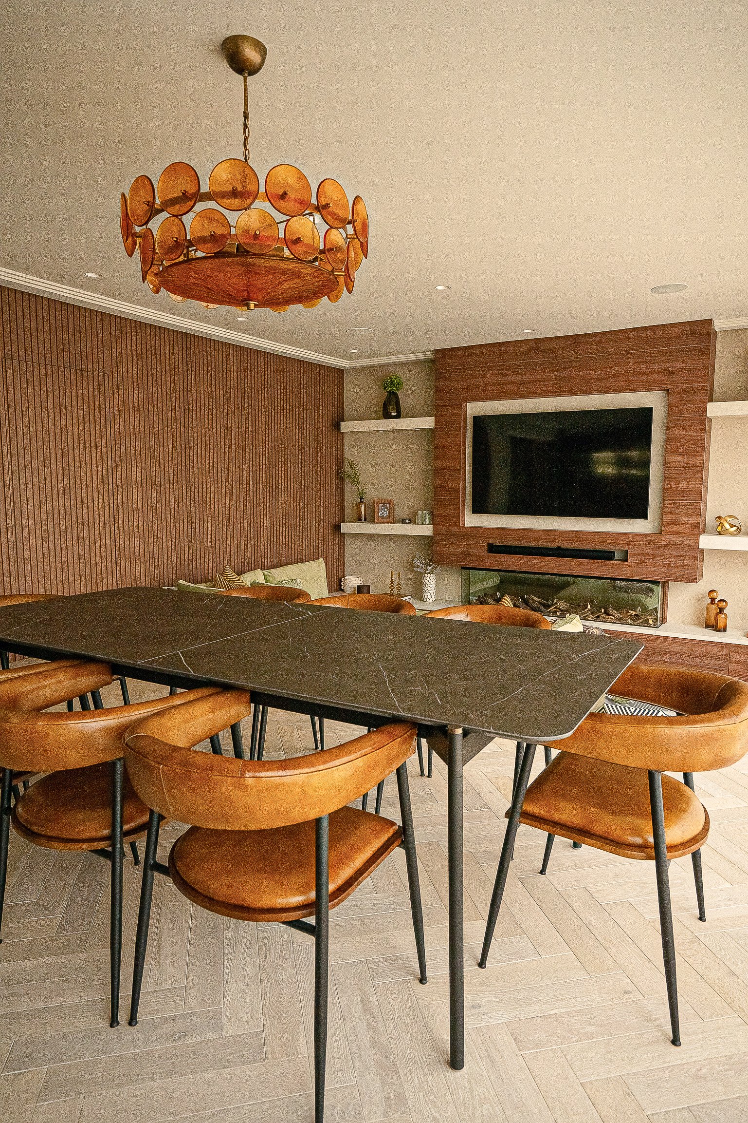 Modern dining room with a black rectangular table, brown leather chairs, and a decorative chandelier. A TV mounted on a wooden accent wall with shelves holding various decor items is in the background.