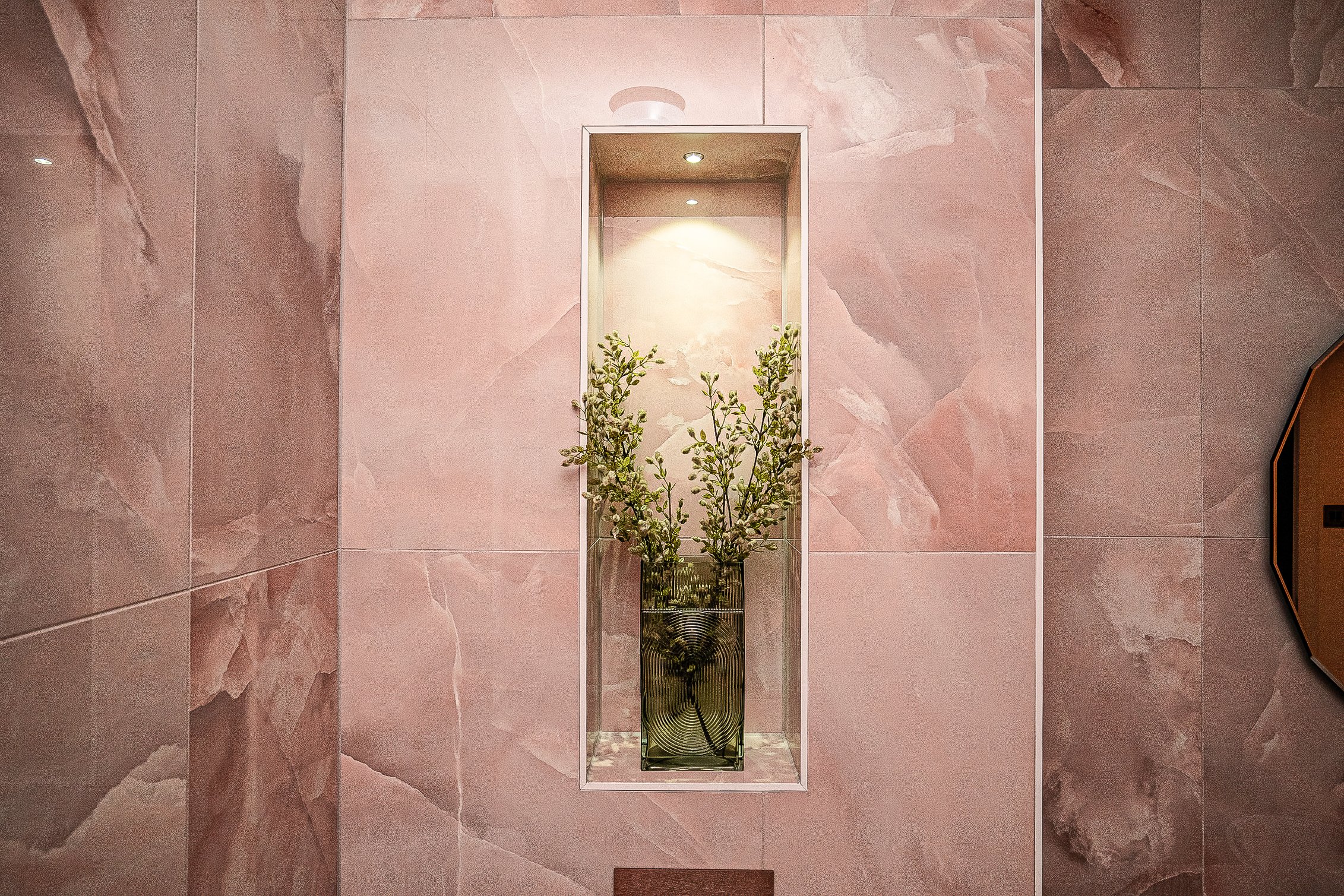 A decorative wall niche with a textured vase holding green branches, set against a pink marble wall with soft lighting.