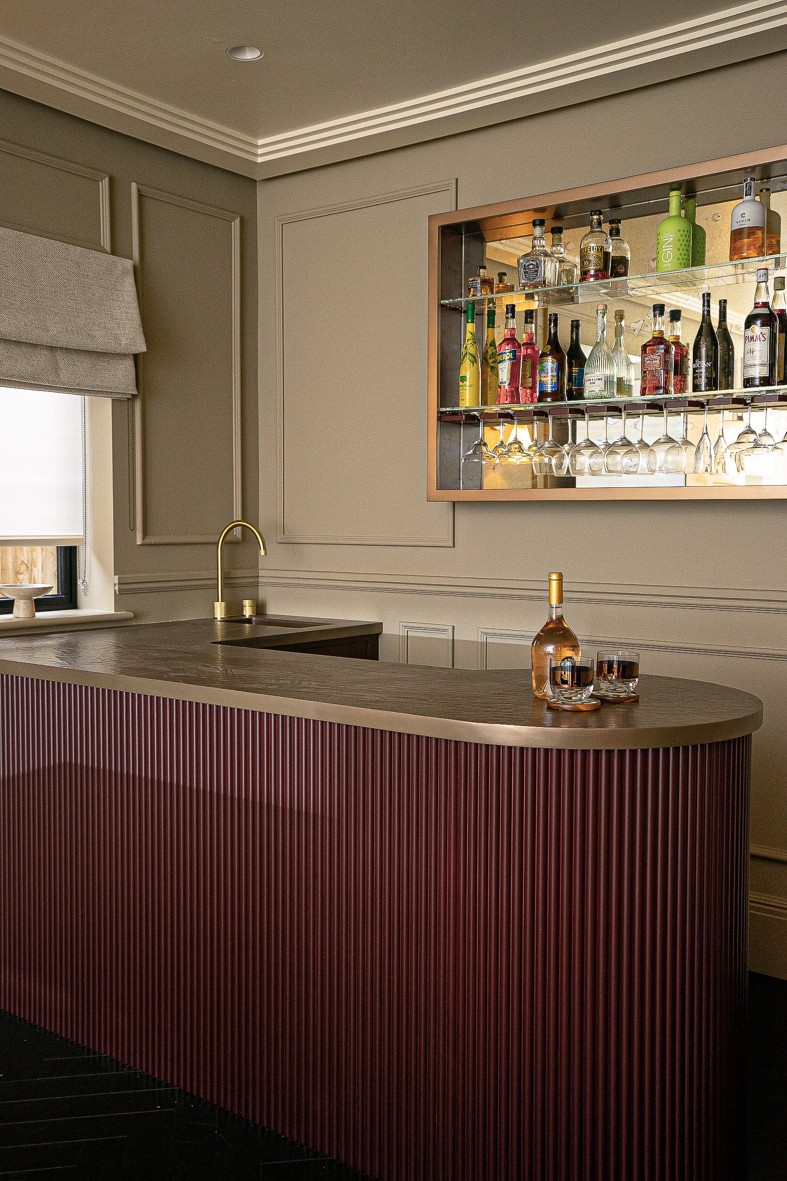 Home bar with wine and liquor bottles on shelves, glassware, wooden counter, gold faucet, and two glasses on the surface.