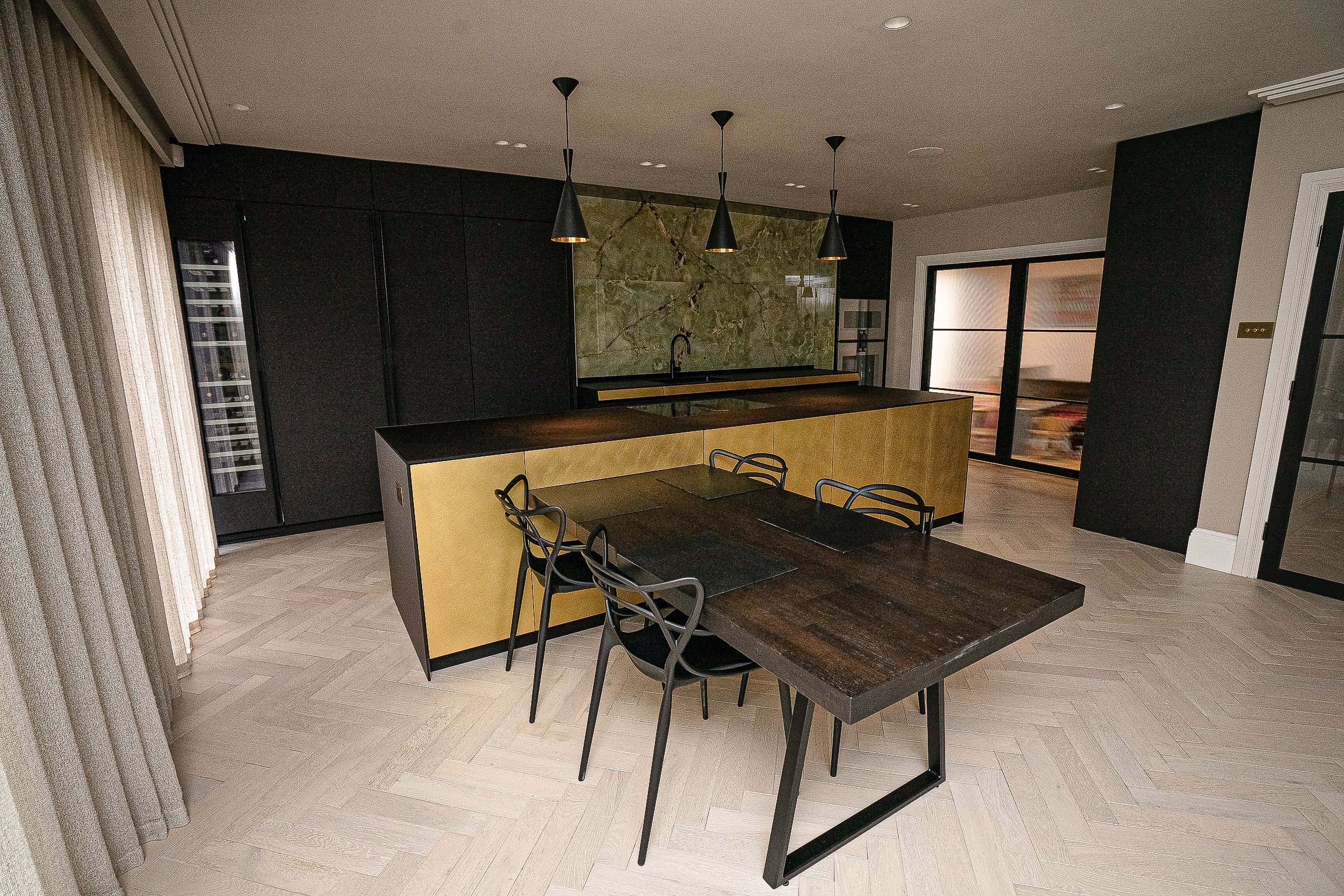 Modern kitchen interior with dark wood table, black chairs, gold island, pendant lights, dark cabinets, and chevron-patterned flooring.