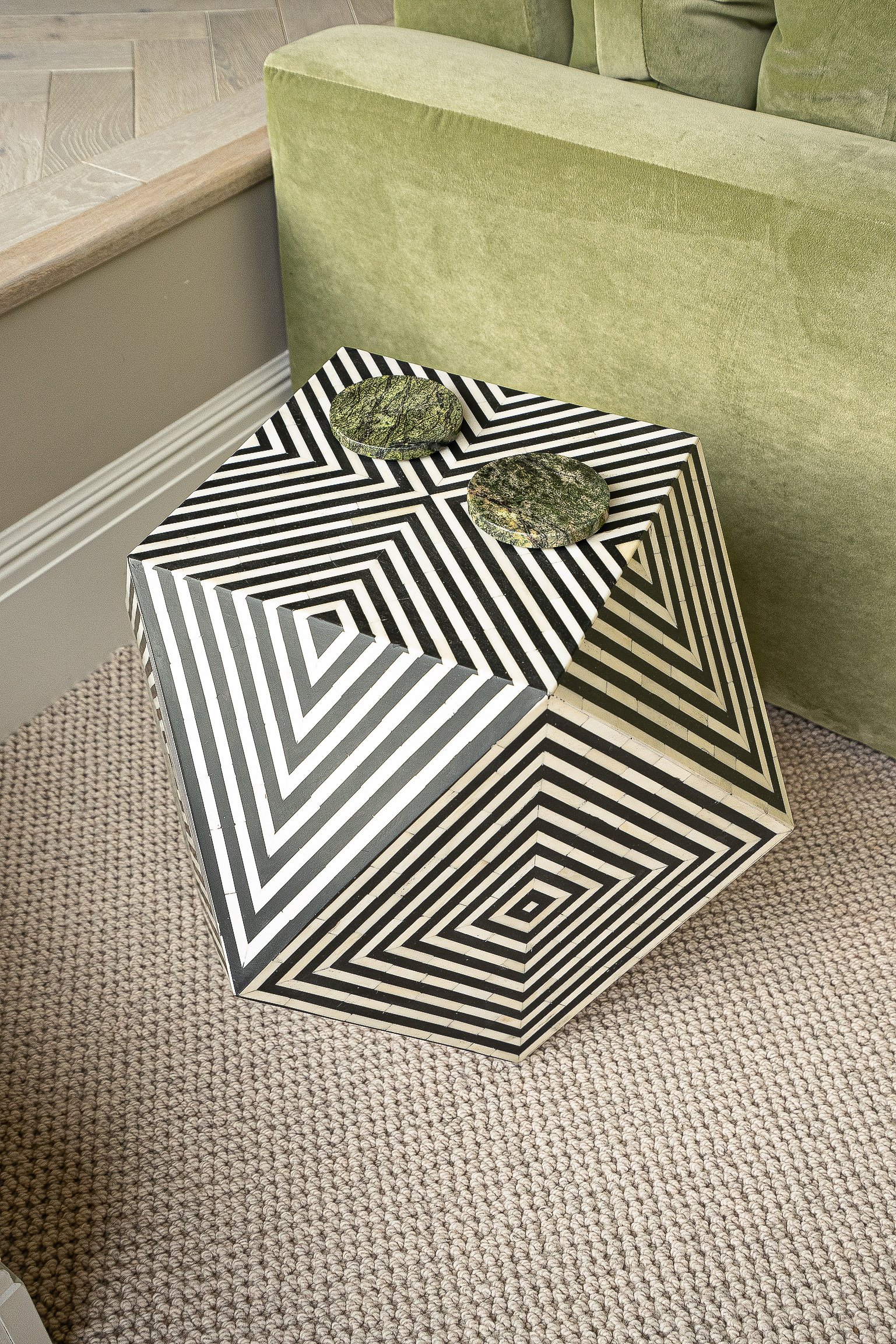 A geometric black and white striped table with two round green coasters on top, placed on a textured carpet next to a green sofa.