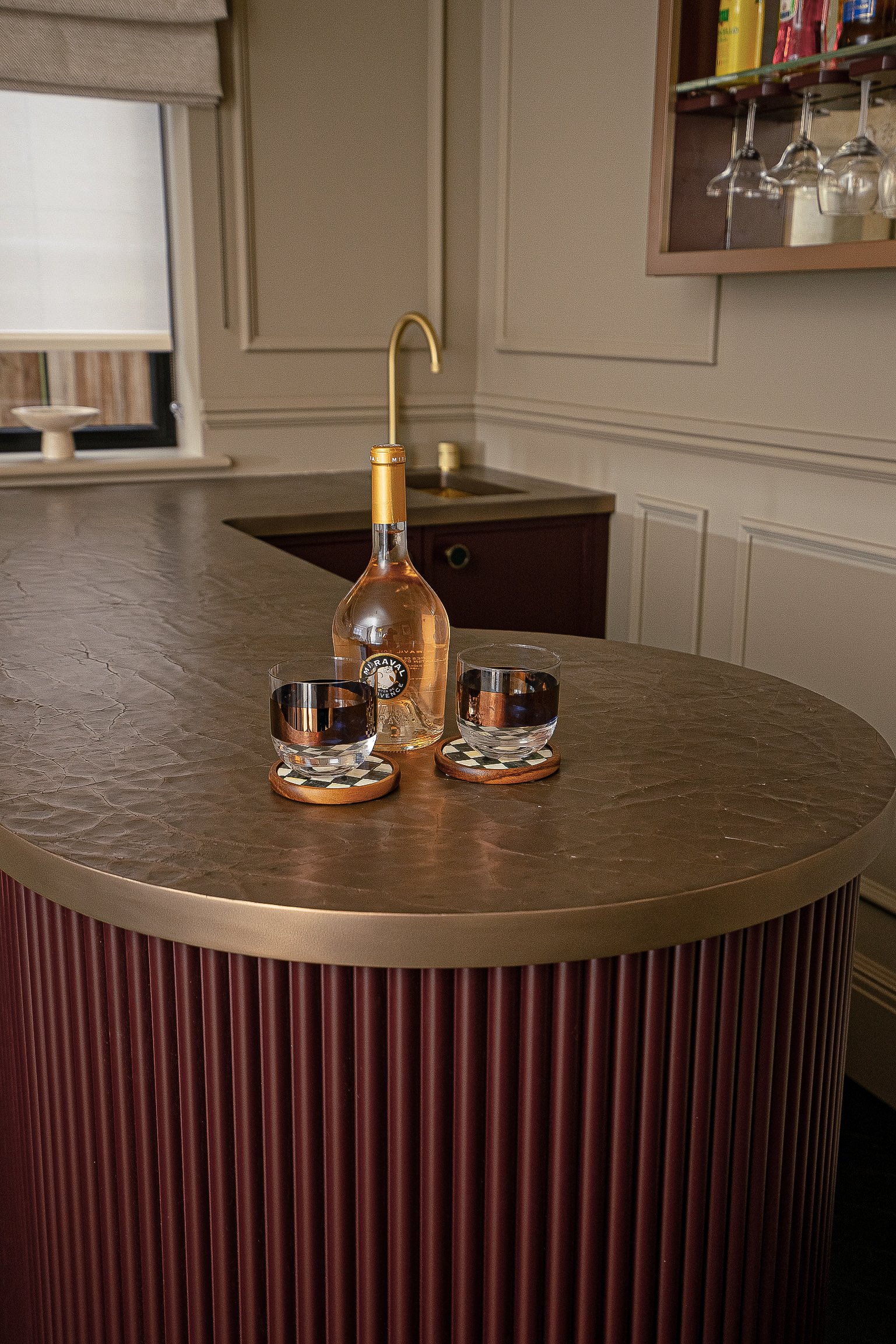 Elegant kitchen with brown countertop featuring a bottle of rosé and two glasses on checkered coasters, gold faucet and shelves in background.