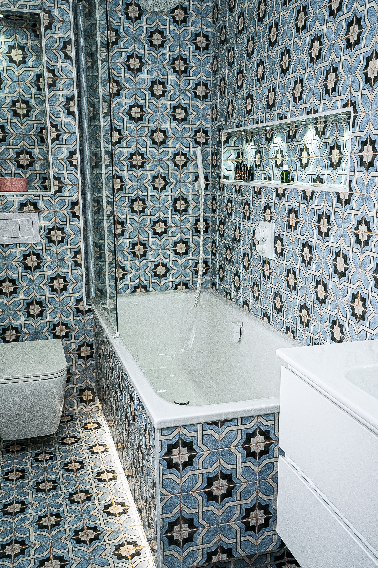 Bathroom with blue geometric patterned tiles, featuring a bathtub with a shower head, toilet, and white sink cabinet.