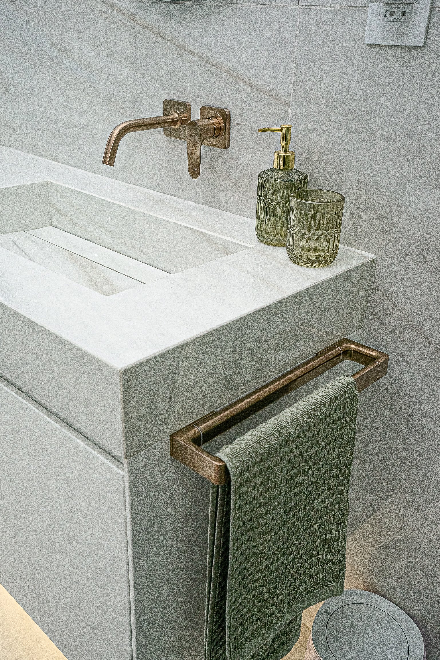Modern bathroom sink with a wall-mounted faucet, green textured towel on a towel rack, green soap dispenser, and a glass on the counter.