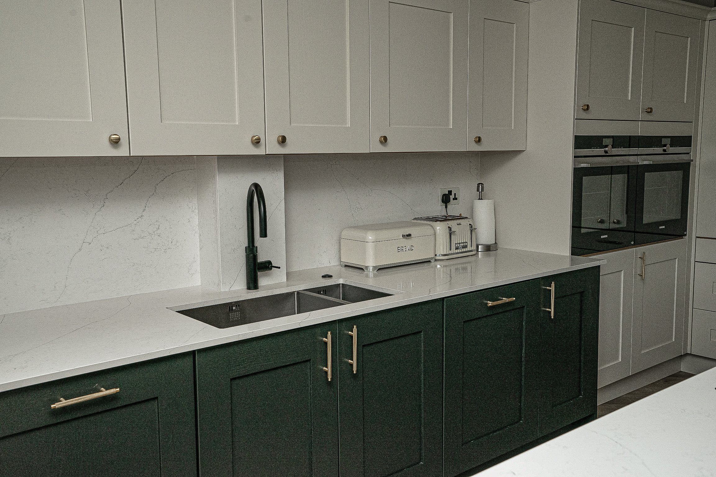 Modern kitchen interior with green and white cabinets, marble countertop, black faucet, double sink, breadbox, toaster, and built-in ovens.