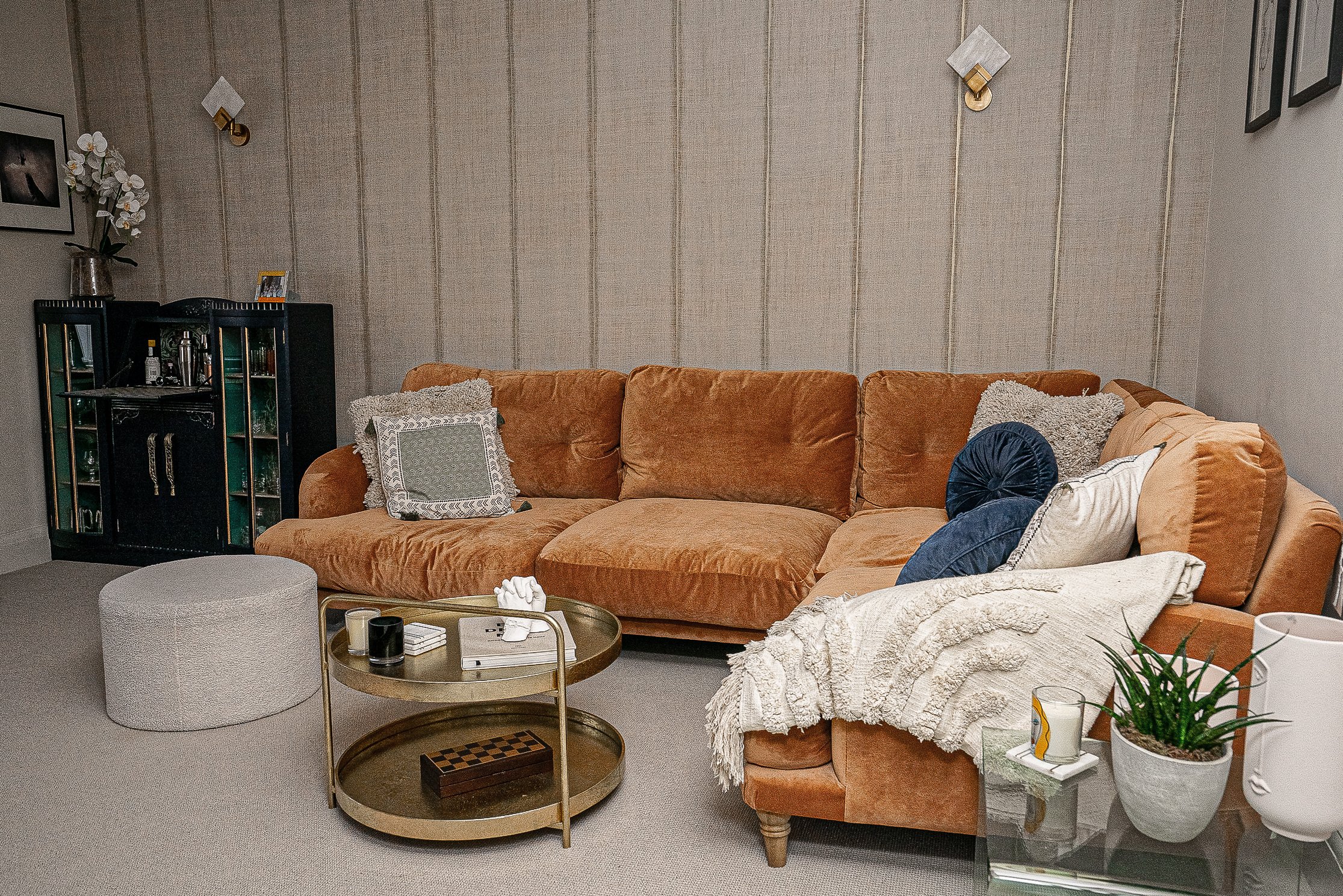 Living room with a brown sectional sofa, decorative pillows, and a round coffee table. A patterned ottoman and potted plant are present. Wall-mounted lights and a small cabinet are in the background. Neutral color scheme.