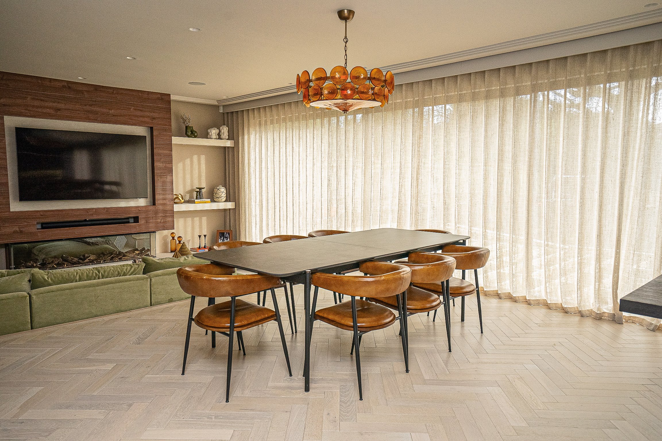 Modern dining room with a wooden table surrounded by eight brown chairs, a chandelier above, and a flat-screen TV on a wooden wall. Large windows with sheer curtains, a green sofa, and built-in shelves with decor are also visible.