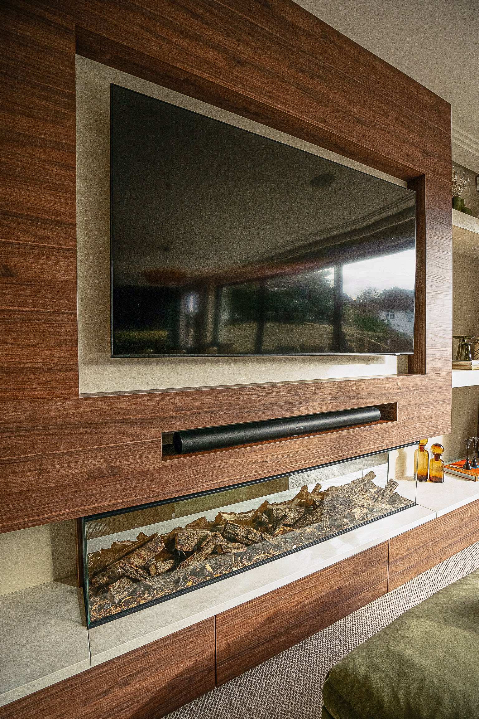 Modern living room with a large mounted flat-screen TV above a soundbar and a glass-enclosed fireplace featuring decorative logs, all set within a wooden panel wall.