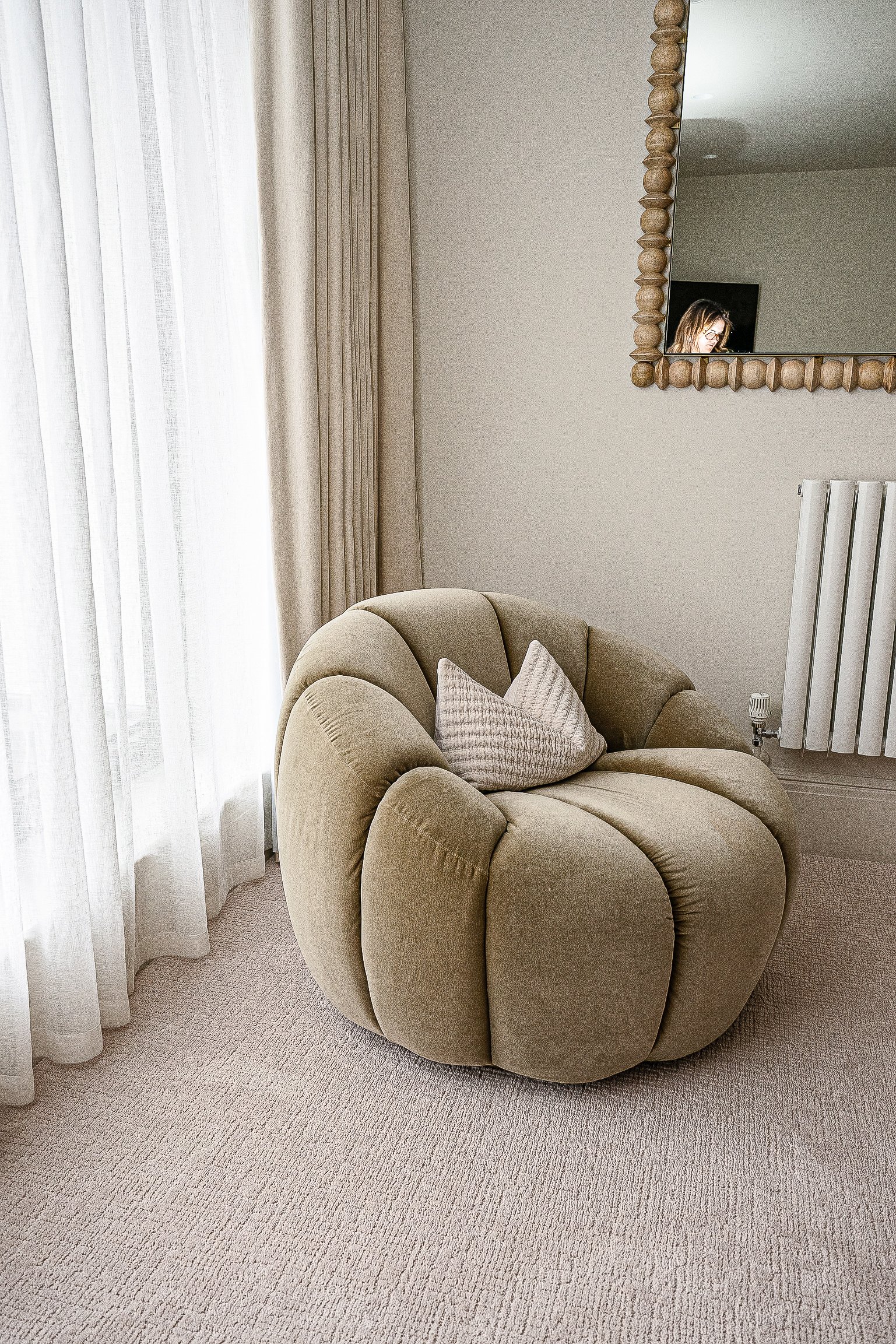 A round, plush beige chair with a decorative pillow in a neutral-toned room featuring a large mirror, radiator, and floor-to-ceiling curtains.