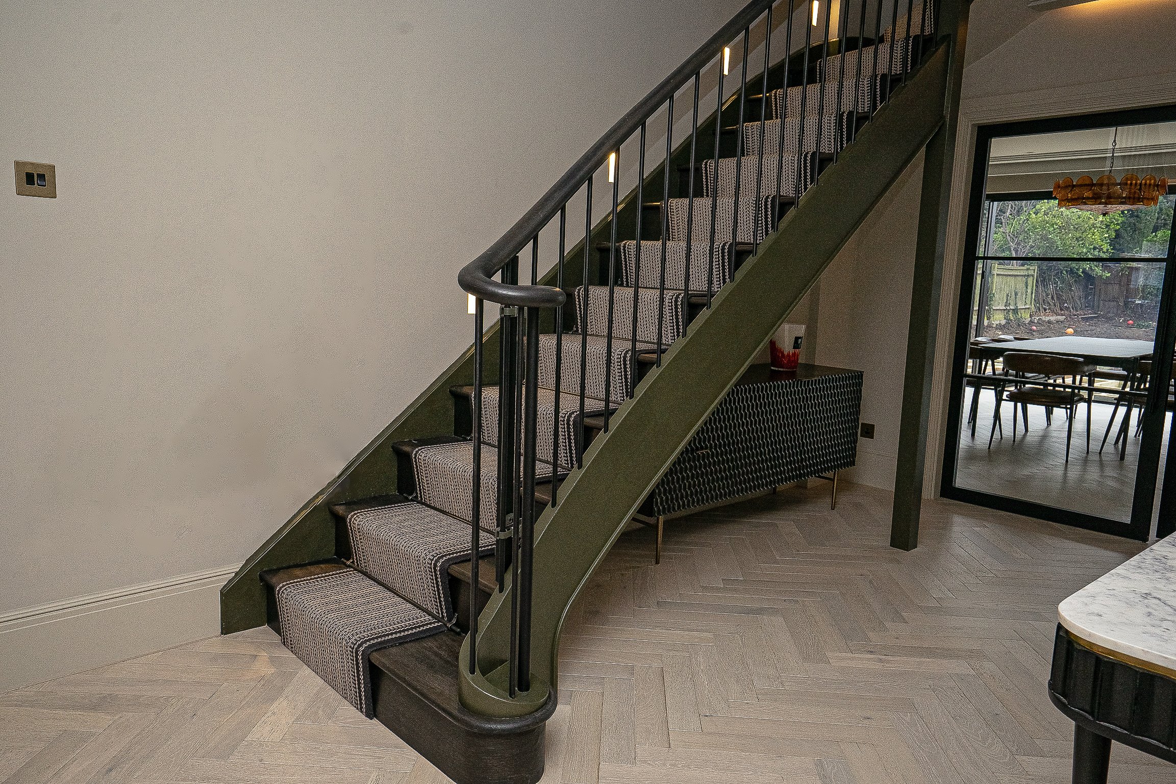 Modern staircase with dark handrails and patterned carpet in a home with light herringbone wood flooring; adjacent room visible through glass door.