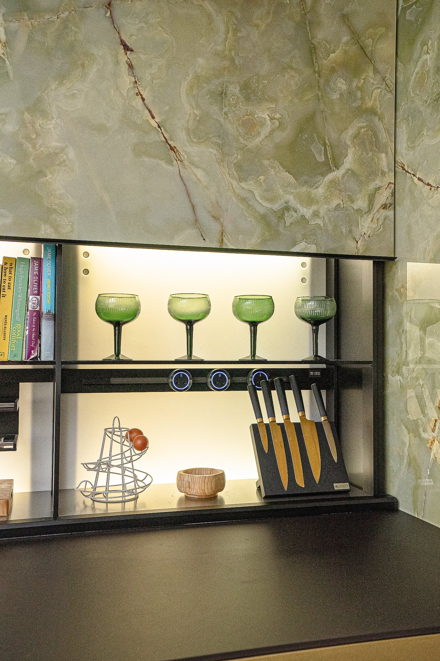 Modern kitchen shelf with green glasses, books, a knife set, and kitchen decor.