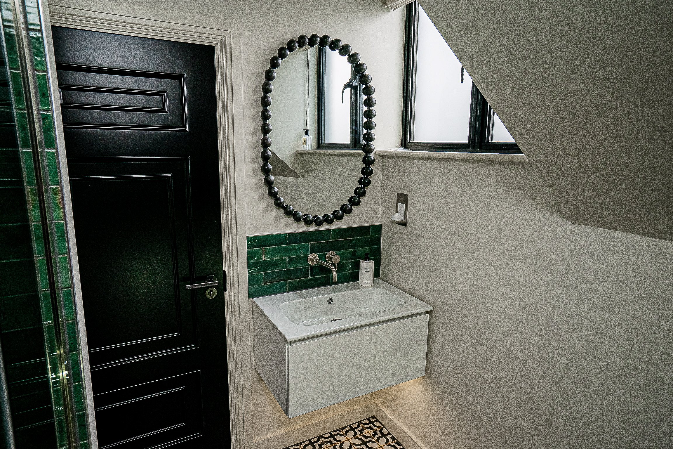 Modern bathroom interior with black door, green tiled wall, white sink, and oval mirror.
