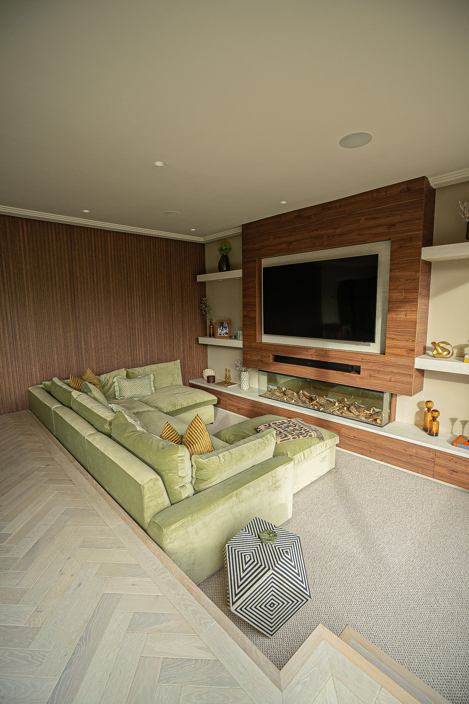 Modern living room with green sectional sofa, large TV on a wooden wall, fireplace, and shelves with decorative items.