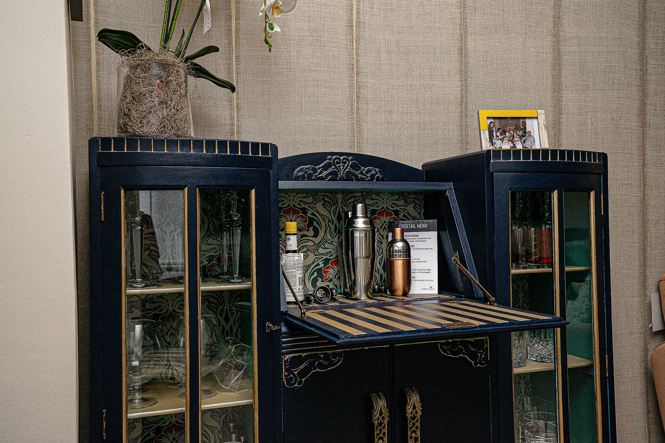 Vintage bar cabinet with glassware, cocktail shakers, and plant on top.