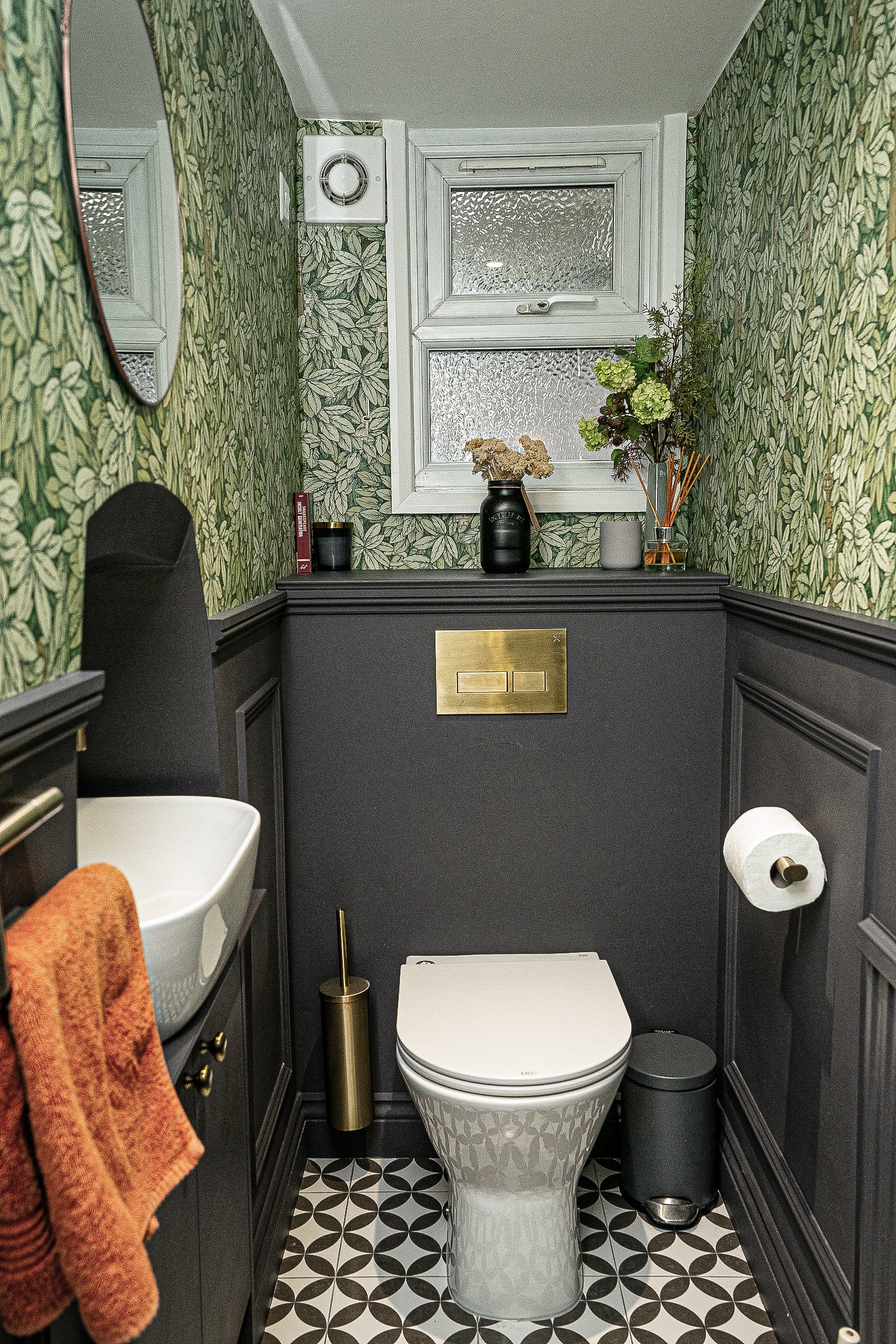Stylish small bathroom with green leaf wallpaper, white toilet, patterned floor tiles, and matte black wainscoting. Features a wall-mounted sink, orange towel, decorative plants, and gold toilet brush holder.