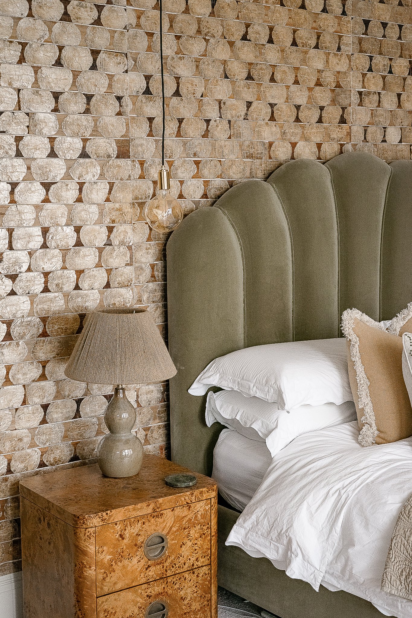 Stylish bedroom interior with green velvet headboard, textured stone wall, wood nightstand, table lamp, and hanging bulb light.