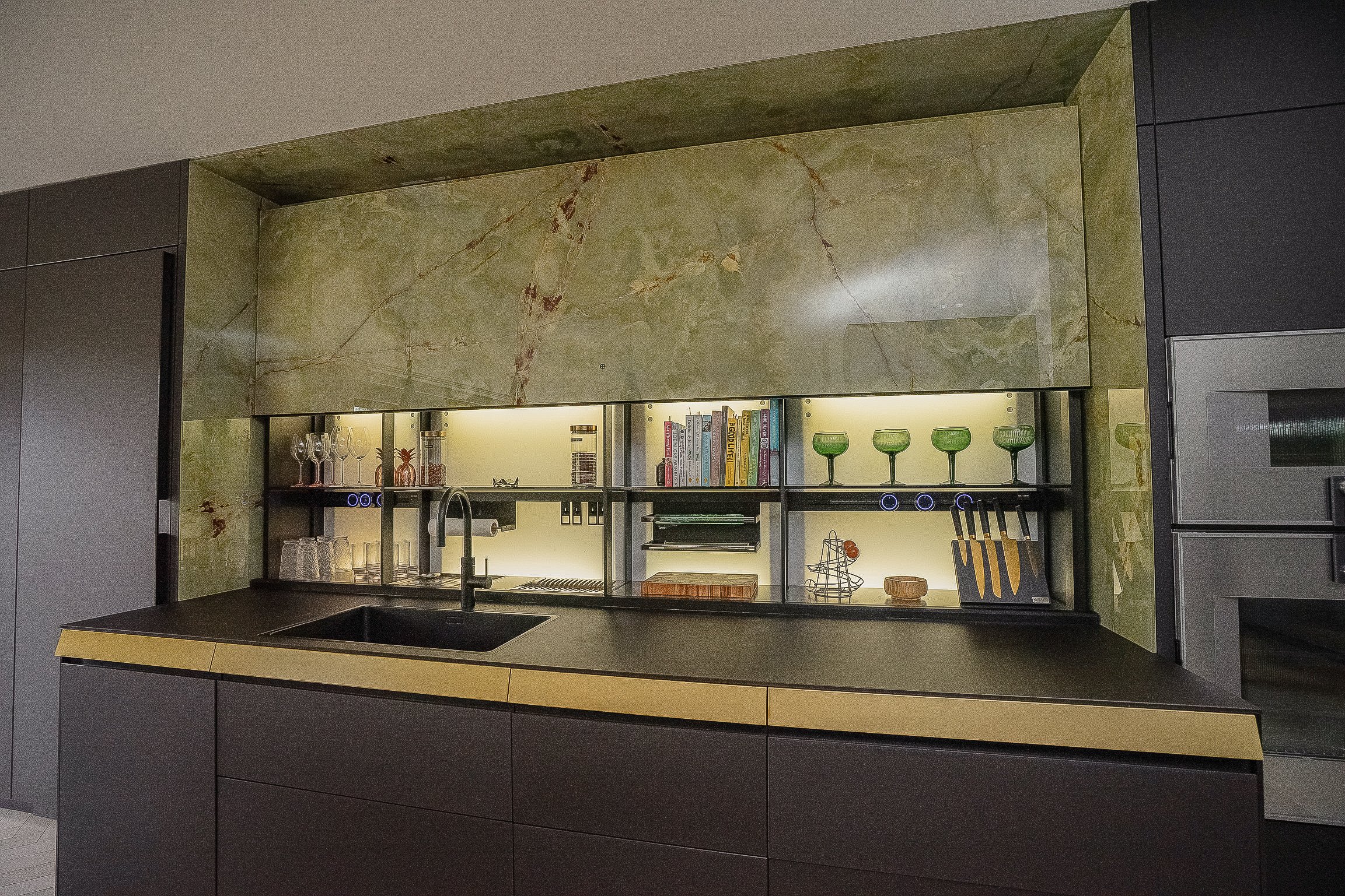 Modern kitchen setup featuring a green marble backsplash, open shelving with glasses, books, and kitchen utensils, black countertop, and sink.