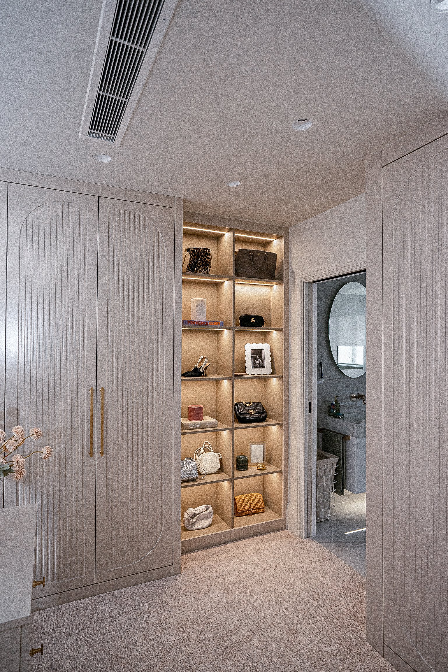 Modern closet with built-in shelves displaying handbags and accessories, alongside a bathroom entrance with a round mirror.