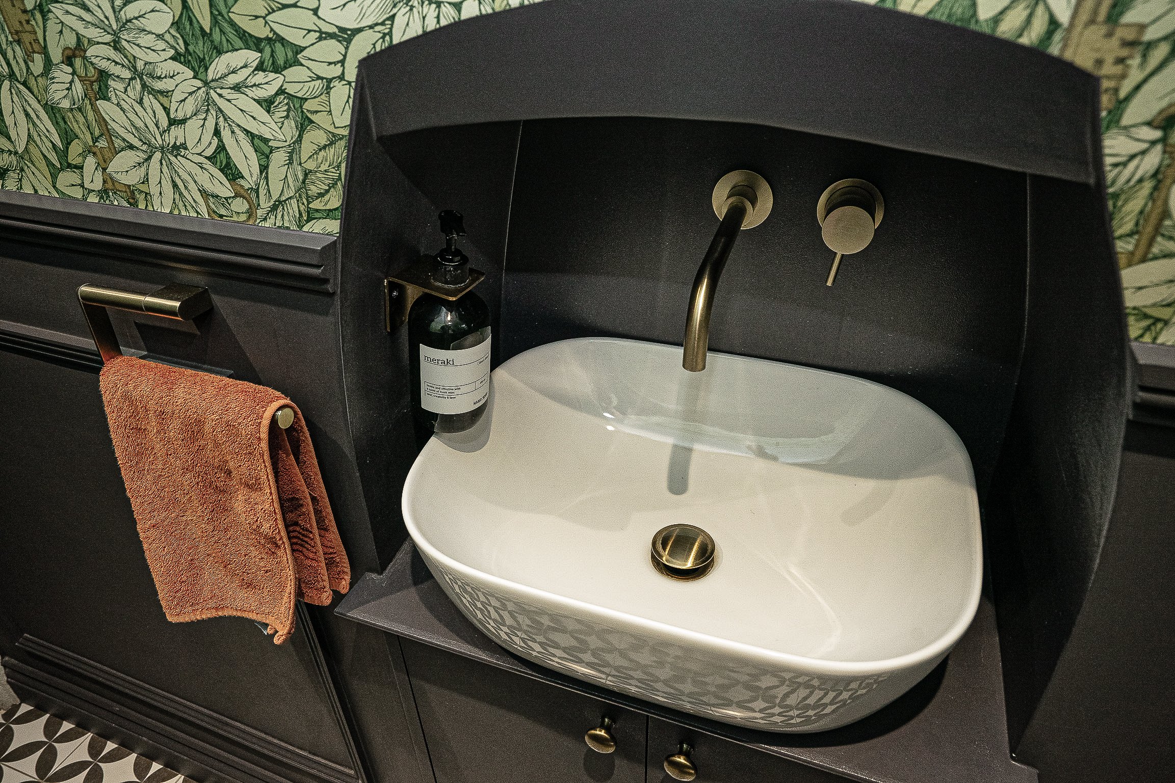 Modern bathroom sink with bronze faucet and handle, green leaf wallpaper, brown towel on towel bar, and a bottle of soap attached to the wall.