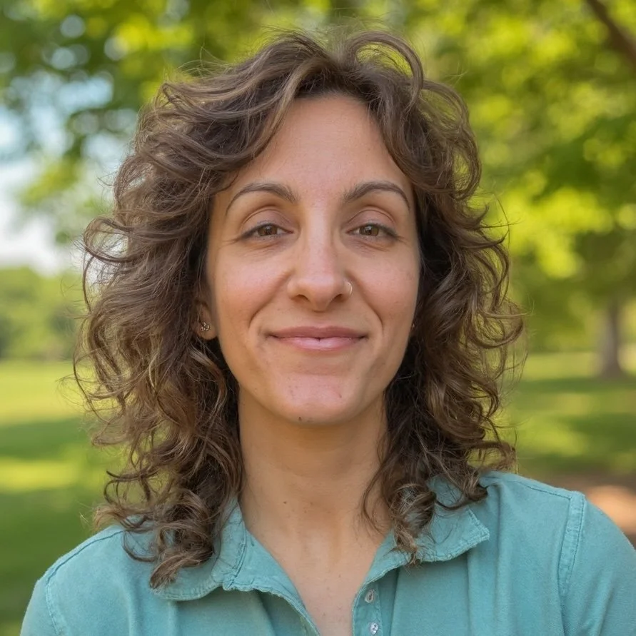 A woman with curly brown hair smiling outdoors with green trees in the background.