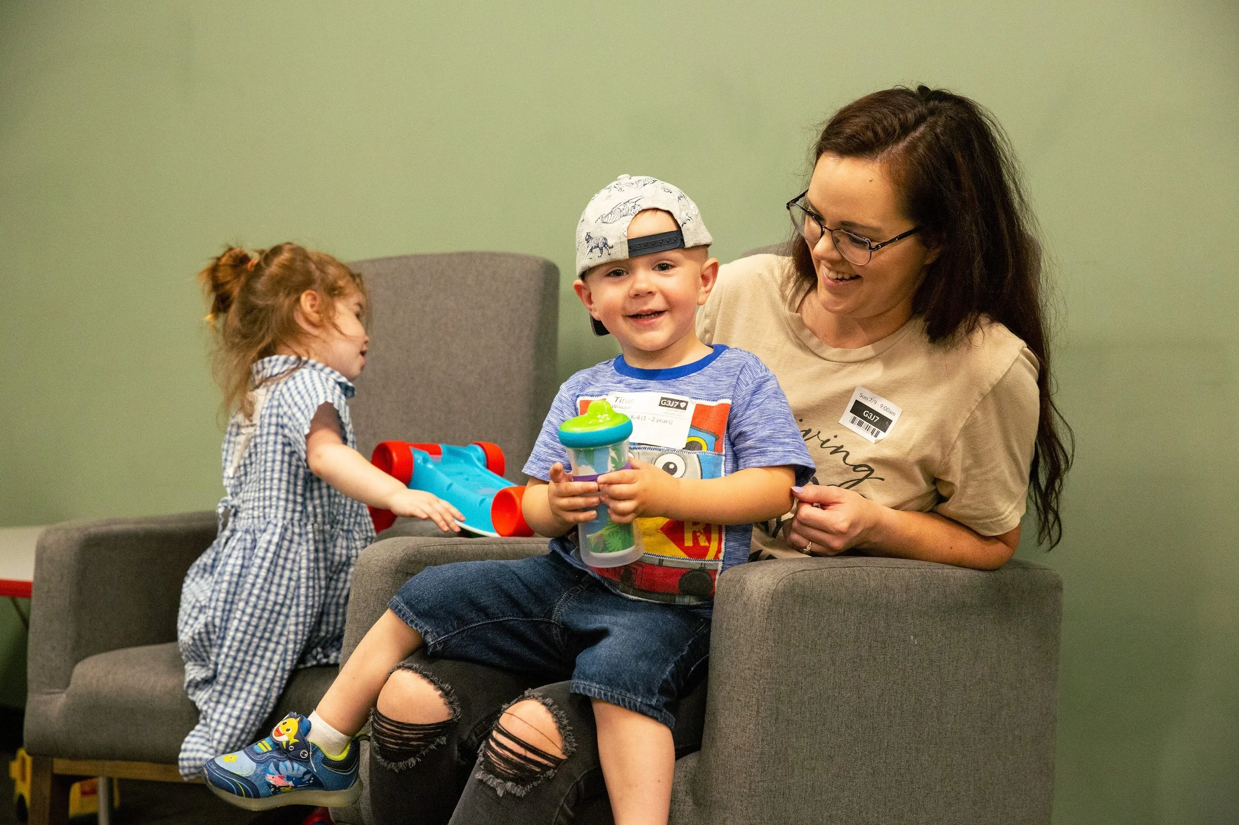 A woman and two children sitting on a gray couch, smiling and playing with toys in a room with a green wall.