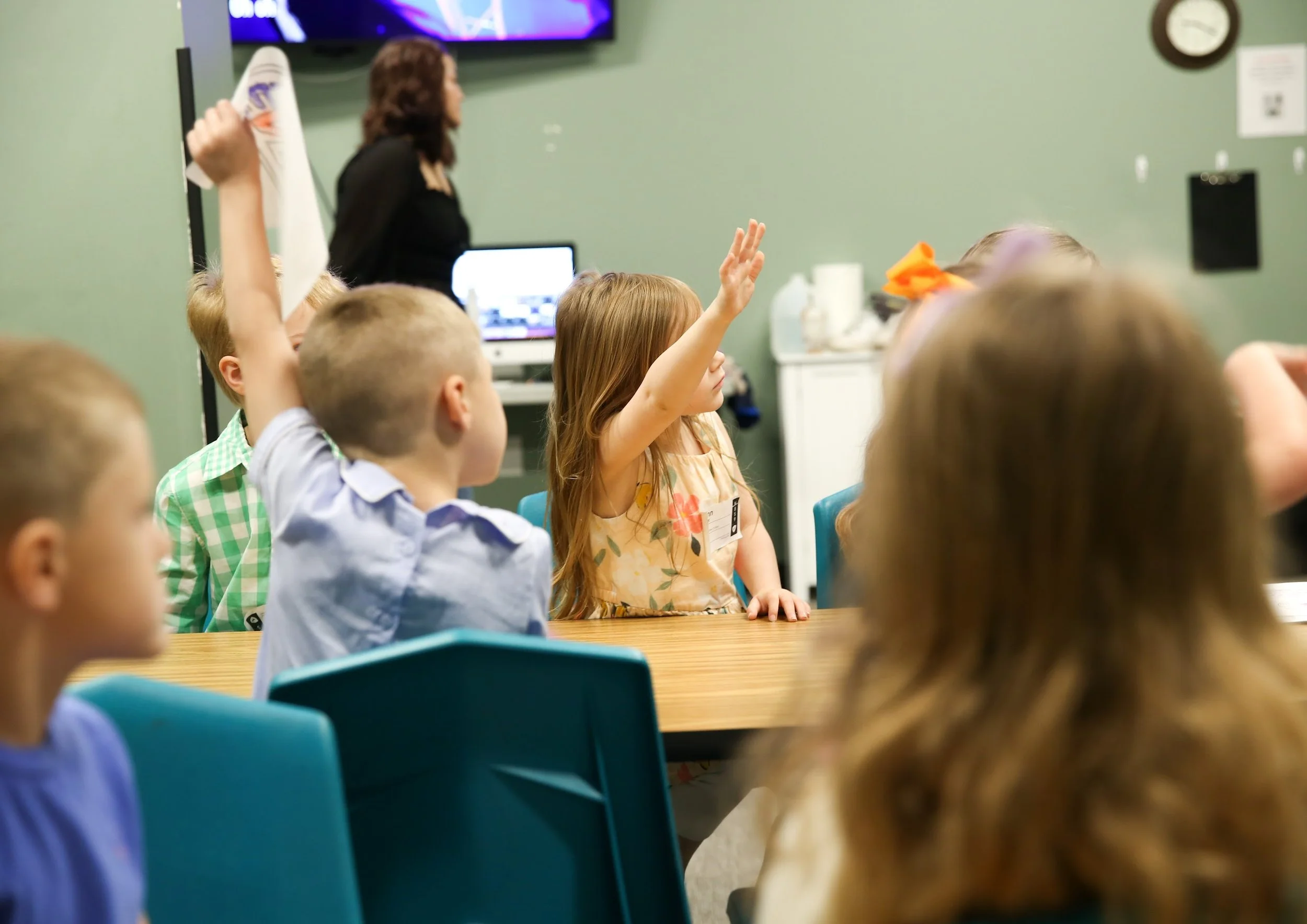 Young girl raising her hand in a classroom with other children and a teacher in the background.