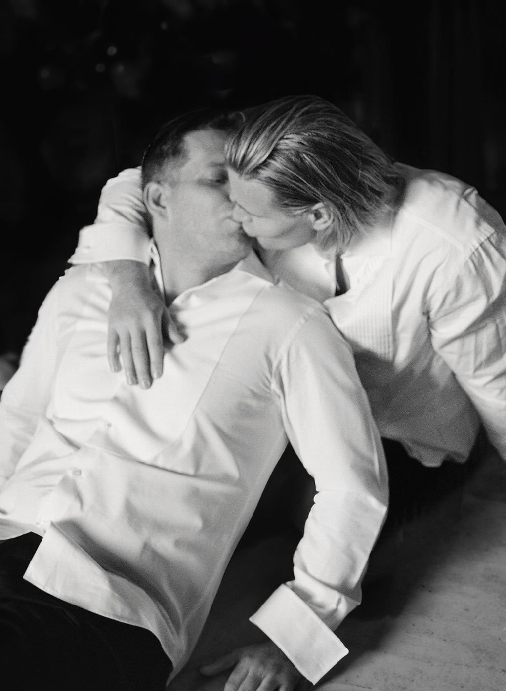 A black and white photo of a gay couple kissing, with the one handsome blond man leaning over the handsome brunete man who is lying on the floor. The photo is taken during their getting ready at the Kulm Hotel in St. Moritz