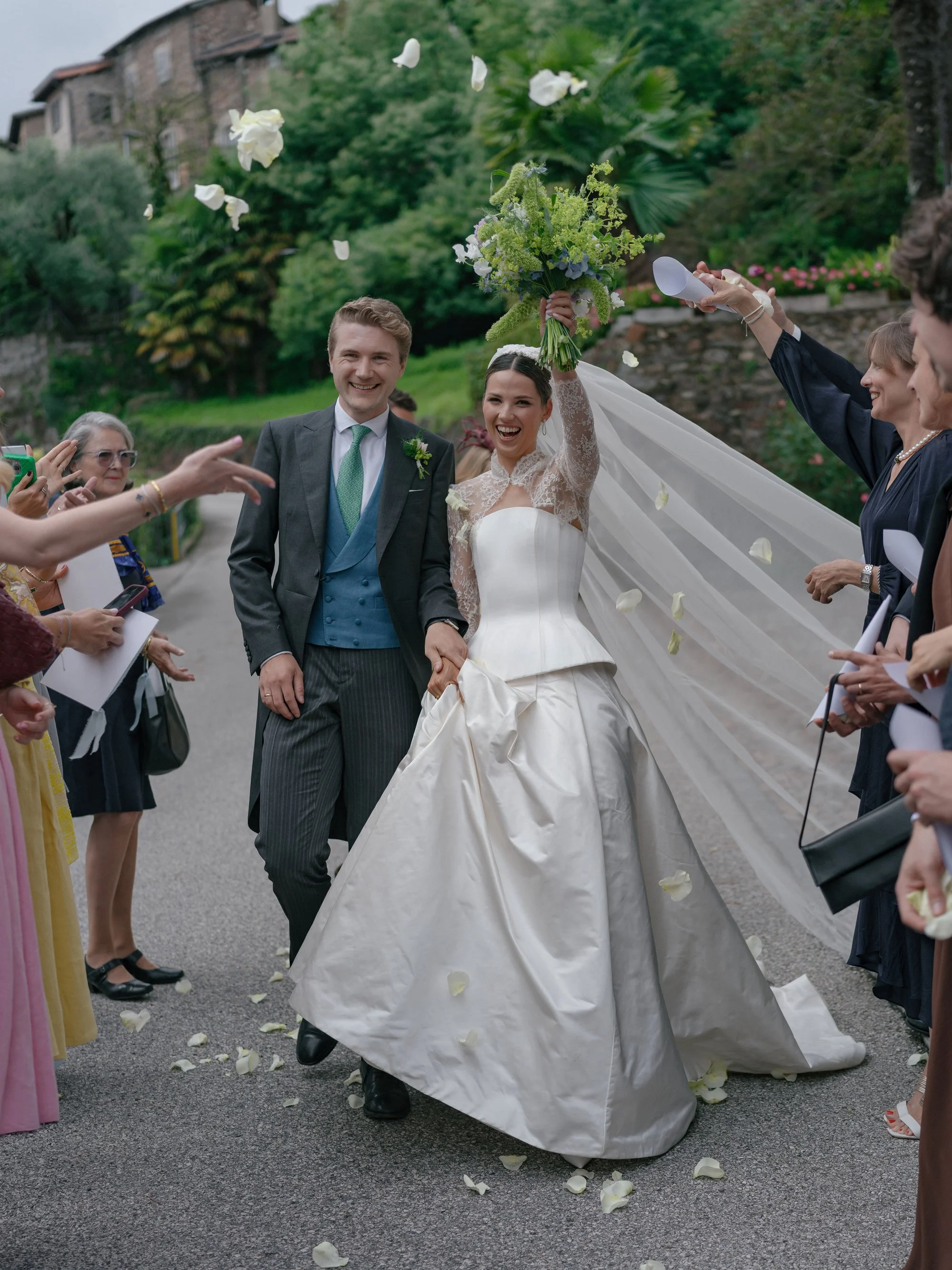 A bride and groom celebrating their wedding surrounded by friends and family, with the bride holding a bouquet and raising her arm while throwing flower petals.
