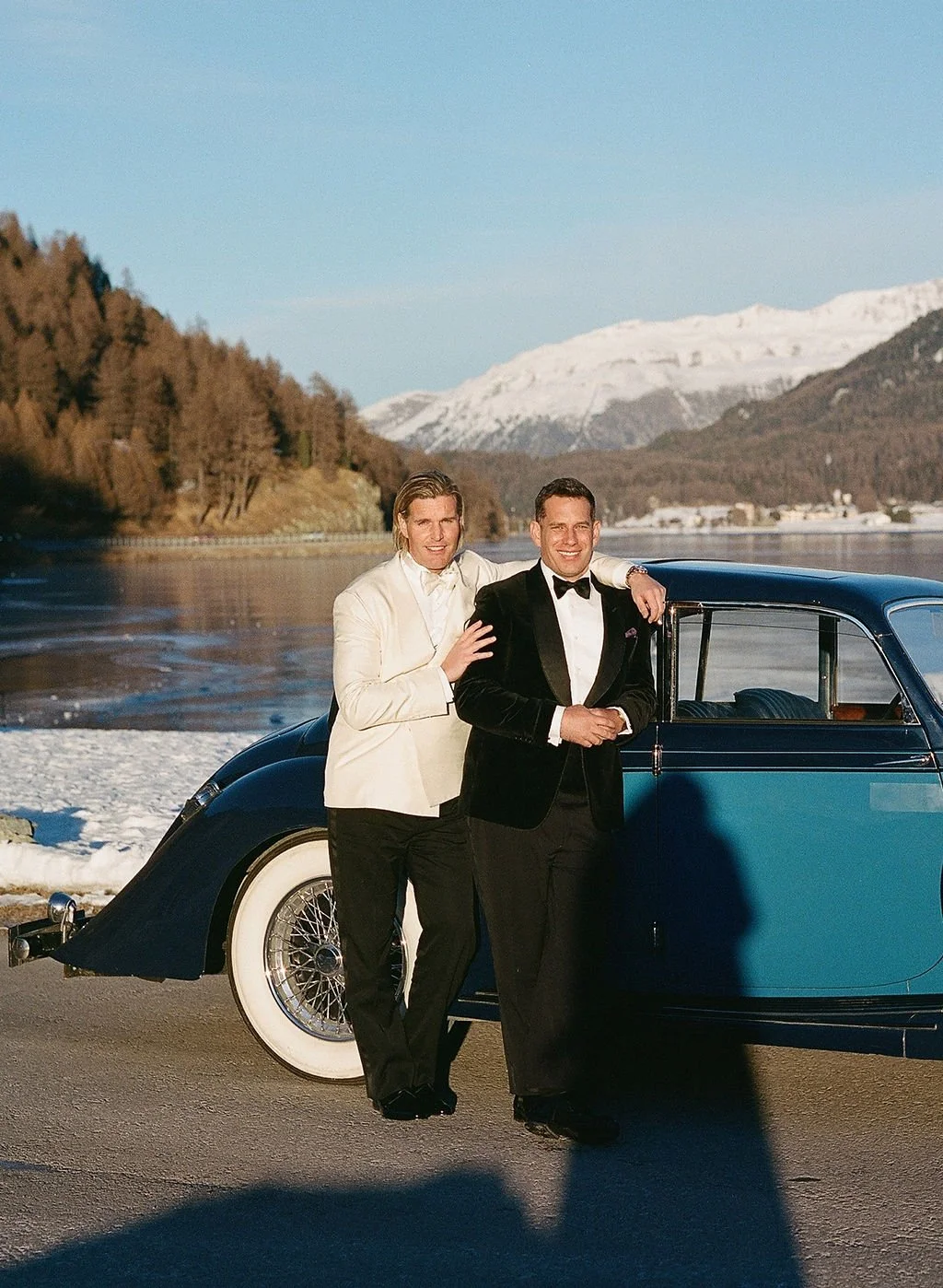 Two men in formal tuxedos standing next to a vintage blue car by a lakeside with snow-capped mountains in the background.