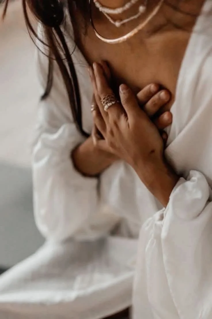 A woman wearing a white blouse and jewelry touching her chest with her hand.