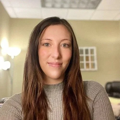 A woman with long brown hair wearing a gray turtleneck sweater taking a selfie in a room with yellow walls and a ceiling light fixture.