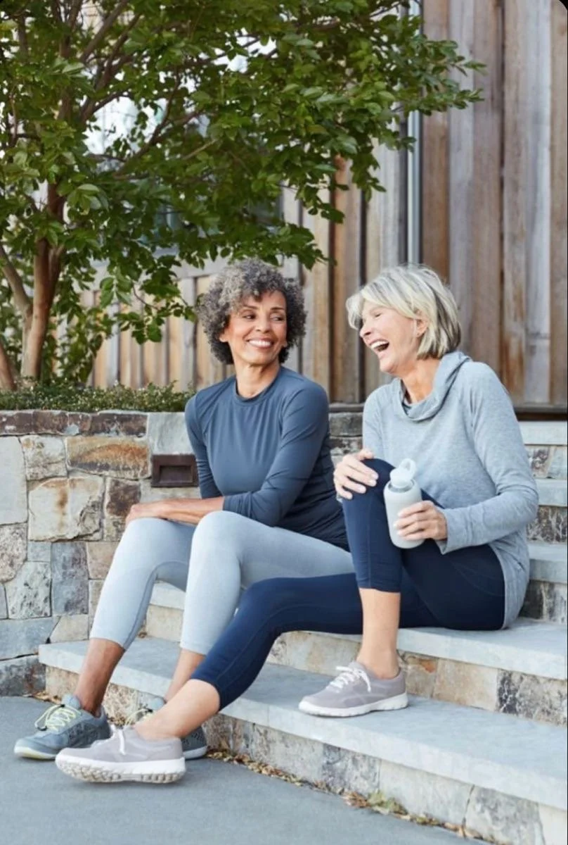 Two women sitting on outdoor steps, smiling and talking, one holding a water bottle. They are dressed in athletic clothing and appear to be enjoying a casual conversation near a tree and wooden fence.