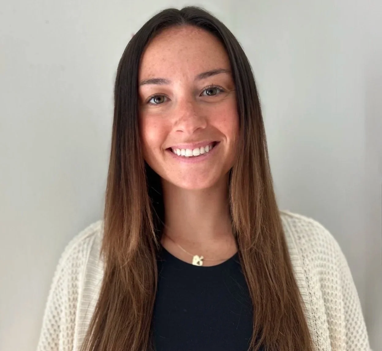 A young woman with long brown hair, green eyes, and freckles, smiling, wearing a black top, a white knitted cardigan, and a necklace with a letter 'K', standing against a plain white wall.