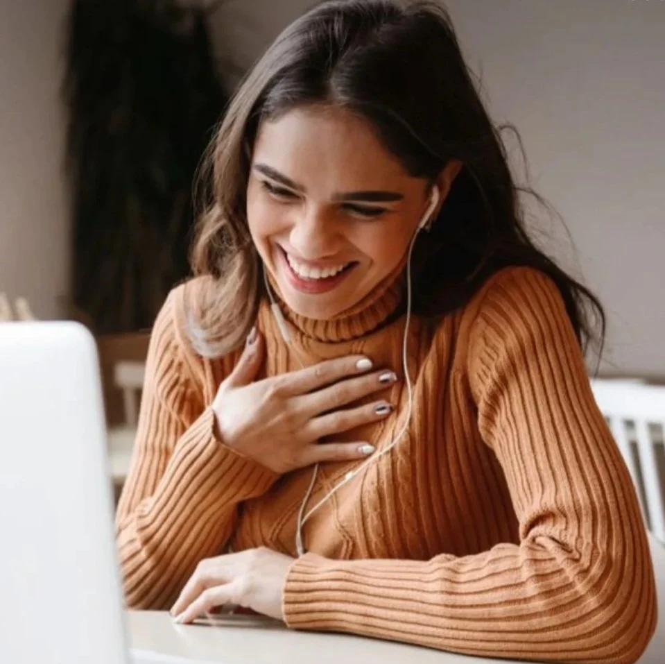 A woman with long dark hair, wearing a brown turtleneck sweater, smiling and seemingly reacting emotionally while looking at her laptop screen, with one hand on her chest and the other on the table.