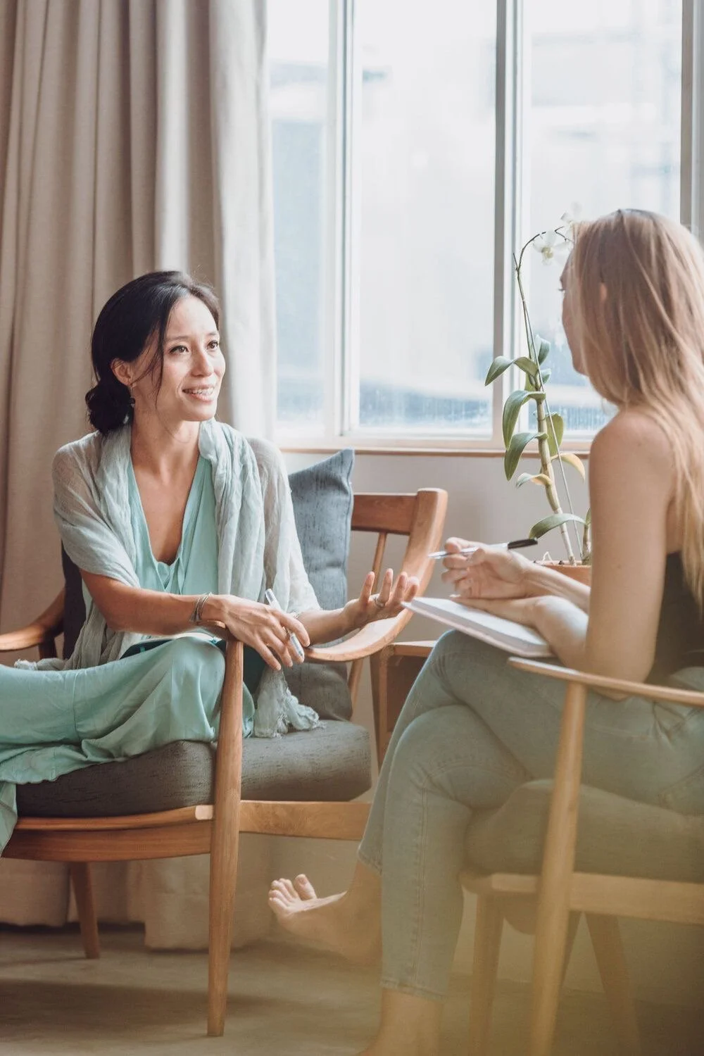 Two women having a conversation in a bright room, one is speaking and gesturing with hands, the other is listening and taking notes. There is a window with curtains and a potted orchid plant.