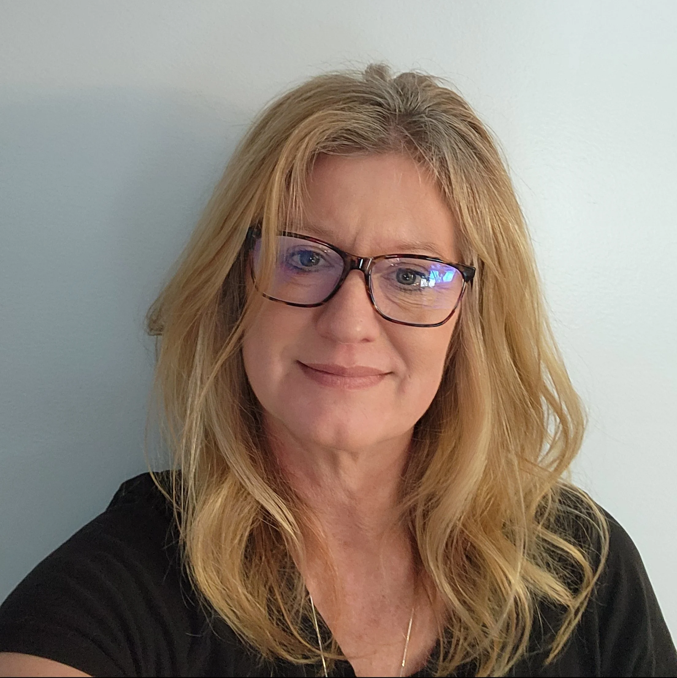 A woman with blonde wavy hair wearing glasses and a black top, standing against a light-colored wall.