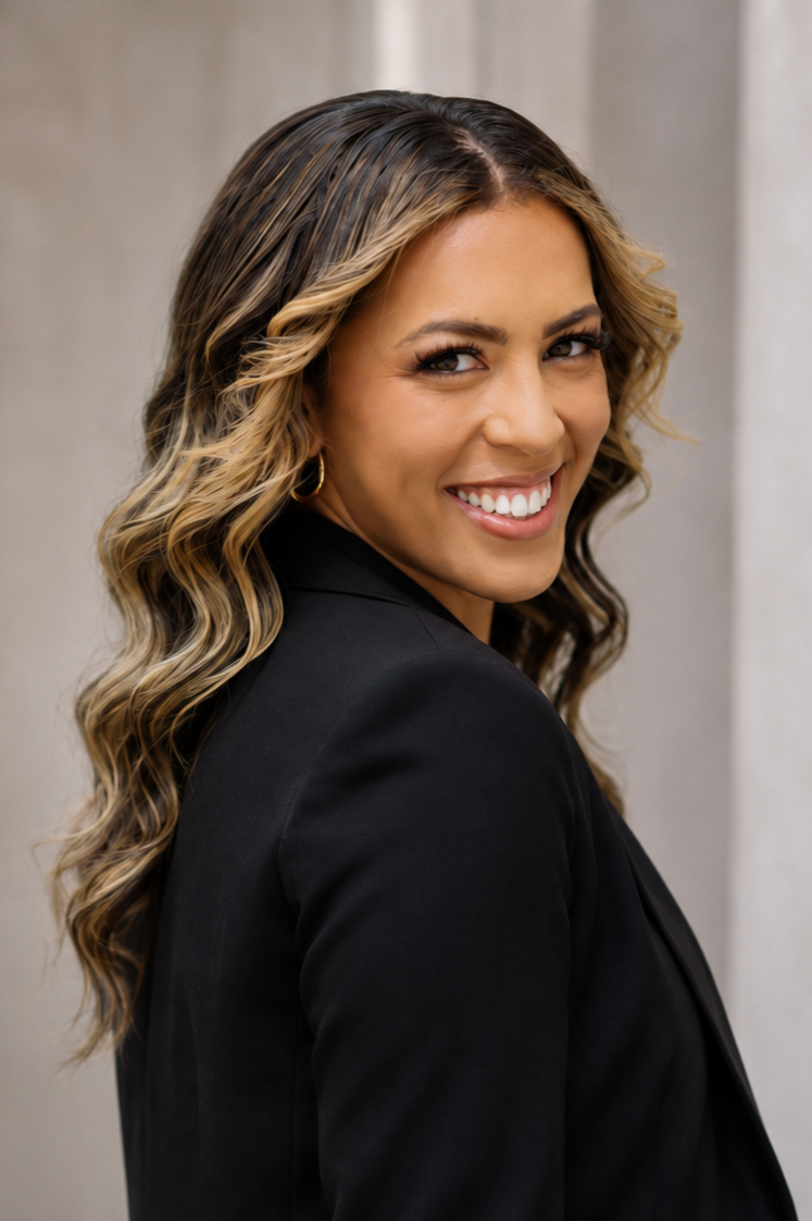 A woman smiling with long, wavy, blonde and brown hair, wearing a black blazer, standing against a neutral background.