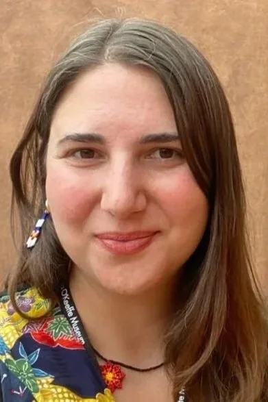 A close-up of a young woman with long brown hair, smiling, wearing a colorful patterned shirt and a black choker, with a plain brown background.