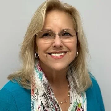 Portrait of a smiling middle-aged woman with blonde hair, wearing glasses, a blue top, and a colorful scarf.