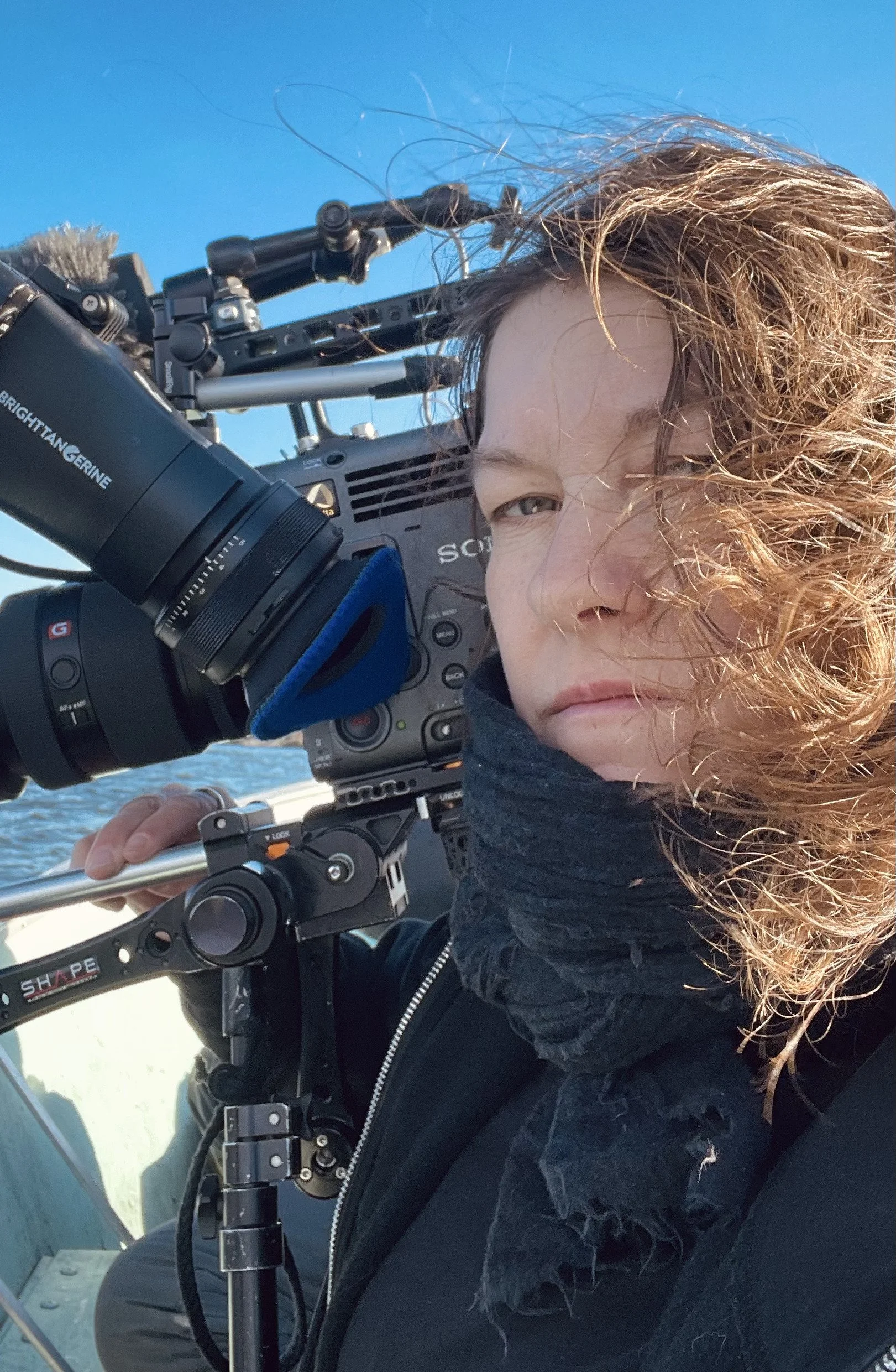 A person with curly hair operating a professional video camera outdoors near water, with a clear blue sky overhead.